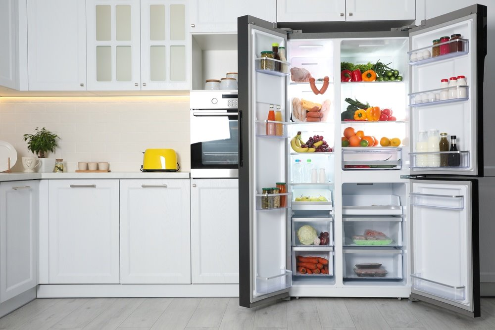 "Open refrigerator in a modern kitchen, filled with various fruits, vegetables, and drinks, with white cabinets and a yellow toaster on the counter."